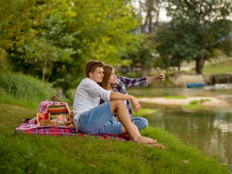 Couple in love taking a selfie by mobile phone while enjoying picnic time drink and food in beautiful nature on the river bank