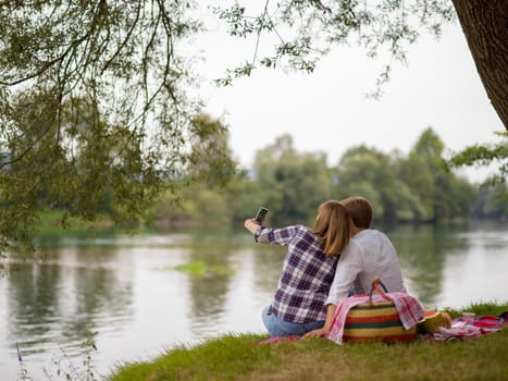 Couple in love taking a selfie by mobile phone while enjoying picnic time drink and food in beautiful nature on the river bank