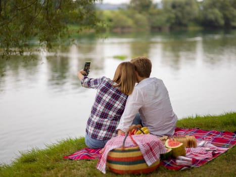 Couple in love taking a selfie by mobile phone while enjoying picnic time drink and food in beautiful nature on the river bank