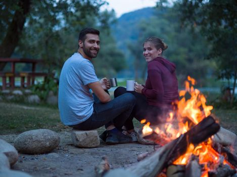 Young couple sitting around the campfire at evening holding a mugs of tea or coffee warming themselves