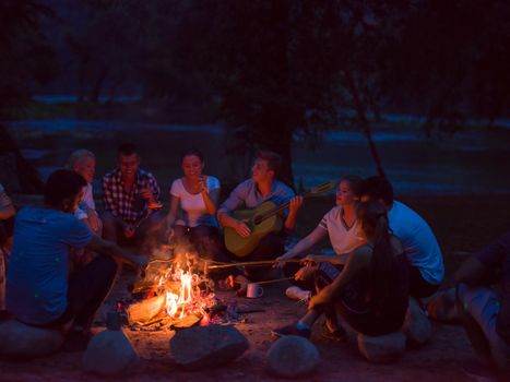 a group of happy young friends relaxing and enjoying  summer evening around campfire on the river bank