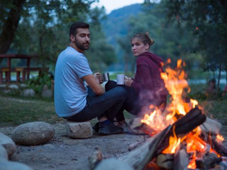 Young couple sitting around the campfire at evening holding a mugs of tea or coffee warming themselves