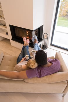 Young romantic multiethnic couple sitting on sofa in front of fireplace at home, looking at each other, talking and drinking coffee