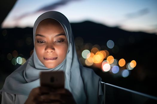 Young Muslim woman wearing scarf veil on urban city  street at night texting on smartphone with bokeh city light in background
