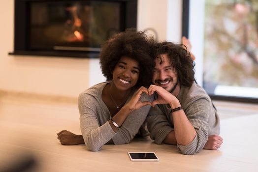 beautiful young multiethnic couple with tablet computer showing a heart with their hands on the floor in front of fireplace at autumn day
