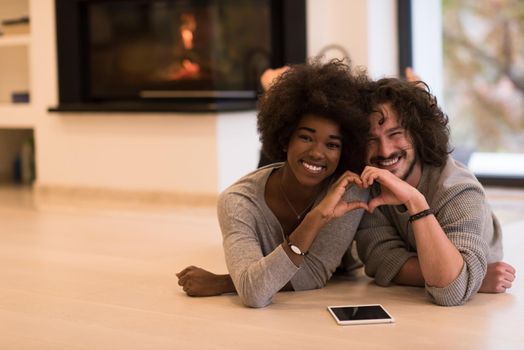 beautiful young multiethnic couple with tablet computer showing a heart with their hands on the floor in front of fireplace at autumn day