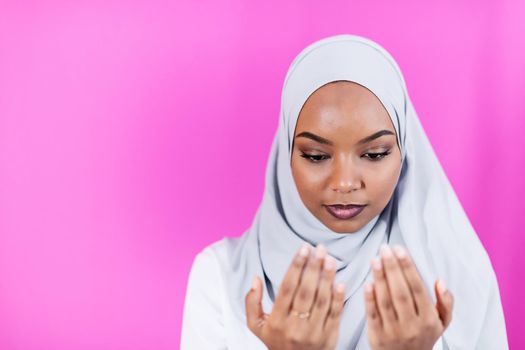 Modern African Muslim woman makes traditional prayer to God, keeps hands in praying gesture, wears traditional white clothes, has serious facial expression, isolated over plastic pink  background