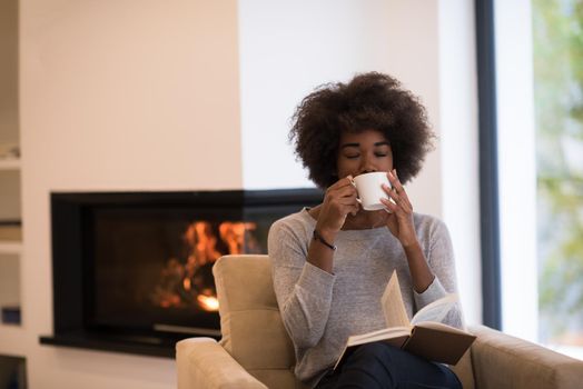 african american woman drinking cup of coffee reading book at fireplace. Young black girl with hot beverage relaxing heating warming up. autumn at home.
