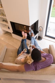 Young romantic multiethnic couple sitting on sofa in front of fireplace at home, looking at each other, talking and drinking coffee