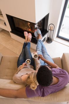 Young romantic multiethnic couple sitting on sofa in front of fireplace at home, looking at each other, talking and drinking coffee