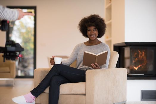 african american woman drinking cup of coffee reading book at fireplace. Young black girl with hot beverage relaxing heating warming up. autumn at home.