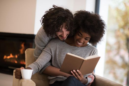 Young beautiful multiethnic couple hugging in front of fireplace at home when reading a book at autumn day