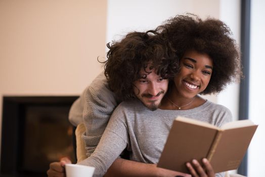 Young beautiful multiethnic couple hugging in front of fireplace at home when reading a book at autumn day