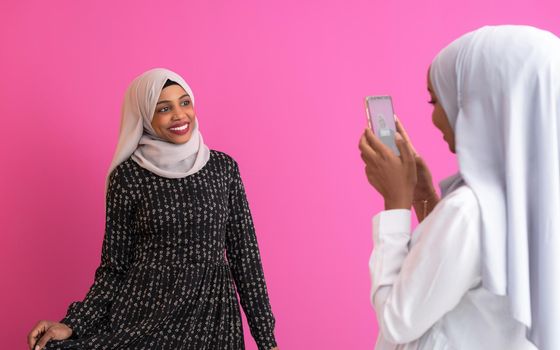 portrait of two diverse african student girls wearing scarf modern muslim clothes  taking self portrait with phone against plastic pink background