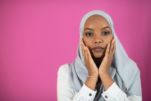 Modern African Muslim woman makes traditional prayer to God, keeps hands in praying gesture, wears traditional white clothes, has serious facial expression, isolated over plastic pink  background