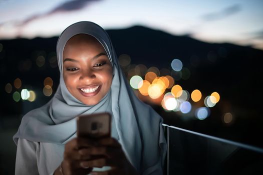Young Muslim woman wearing scarf veil on urban city  street at night texting on smartphone with bokeh city light in background