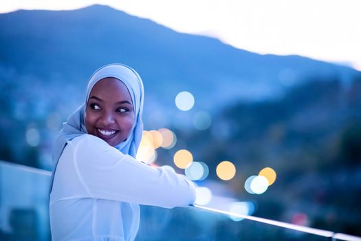African  modern Muslim woman in night  at balcony  smiling at camera with city bokeh lights in background