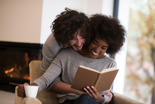 Young beautiful multiethnic couple hugging in front of fireplace at home when reading a book at autumn day