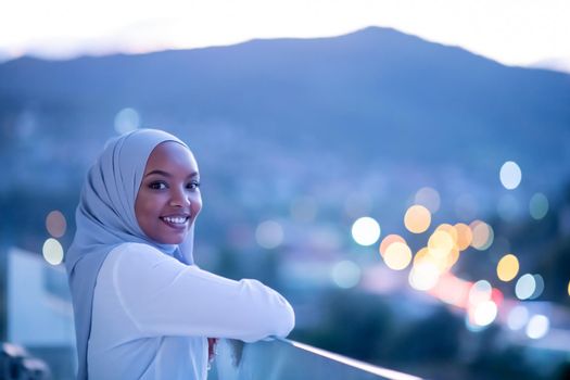 African  modern Muslim woman in night  at balcony  smiling at camera with city bokeh lights in background
