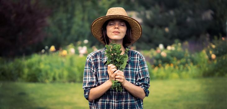 A young girl gardener in a straw hat holds a bouquet of harvested fresh mint and inhales its wonderful menthol scent, a woman is harvesting in the garden.