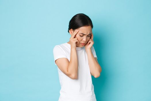 Side view of bothered tired asian girl grimacing, touching head and complaining on headache, having pain in head, migraine of feeling dizzy, standing blue background sick.