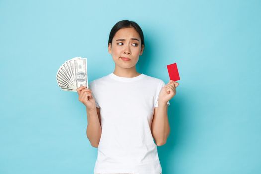 Shopping, money and finance concept. Indecisive and confused attractive asian girl cant decide whats better, credit card or cash, looking perplexed, standing thoughtful over blue background.