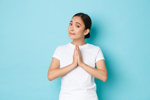 Silly cute asian girl need help, holding hands together in pleading gesture, begging for favour, lend money, standing blue background supplicating or apologizing with gloomy smile.