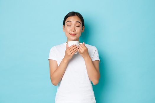 Portrait of delighted dreamy, pretty asian girl in white t-shirt, close eyes and smelling coffee, enjoying morning routine, sipping cappuccino from mug, standing light blue background.