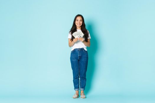 Fashion, beauty and lifestyle concept. Full-length of smiling young asian woman showing dollars, earn money and looking satisfied, winning lottery, standing with cash over light blue background.