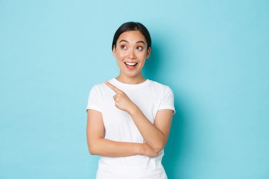 Lifestyle, beauty and shopping concept. Excited and amused beautiful asian girl in white t-shirt pointing upper left corner and smiling thrilled with awesome advertisement promo, blue background.