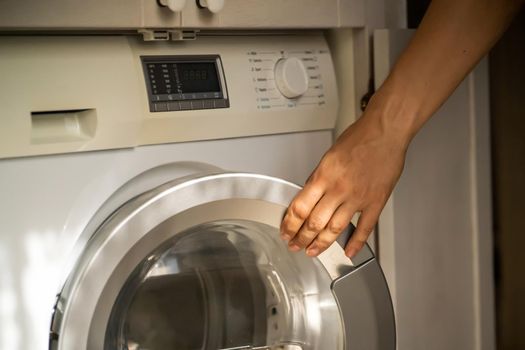 A male hand opens the door of a new washing machine in his modern, bright bathroom, close-up. The man washes clothes in the laundry.