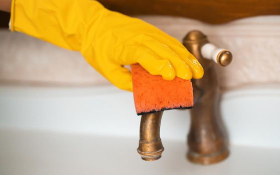 A woman in rubber gloves is cleaning the house, creating cleanliness and disinfection, wiping the faucet and wash basin in the bathroom with a sponge.