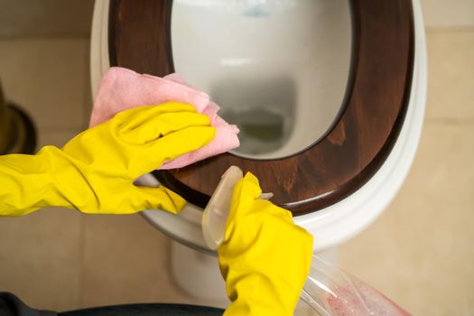 Female hands in yellow rubber gloves are holding a rag and wiping the toilet with detergent and disinfectant. The woman is cleaning the bathroom.