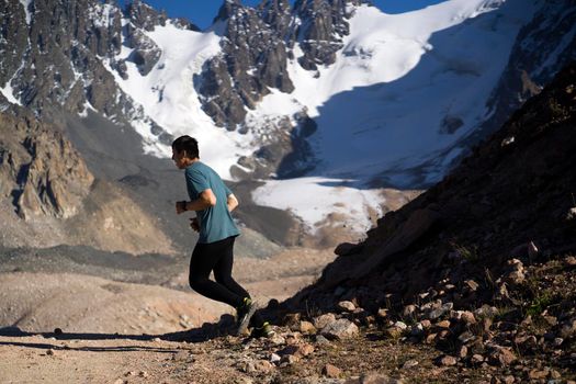 A young man runs along a trail in the snow-capped mountains, doing outdoor workout. The runner warms up, prepares for the race, leads a healthy active lifestyle.