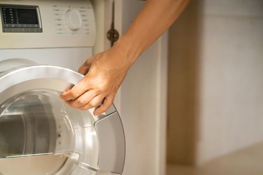 A male hand opens the door of a new washing machine in his modern, bright bathroom, close-up. The man washes clothes in the laundry.