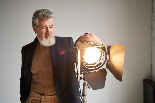 Portrait of handsome bearded middle aged man dressed elegantly standing next to studio spotlight while posing for camera over white background. Fashion photoshoot, style concept