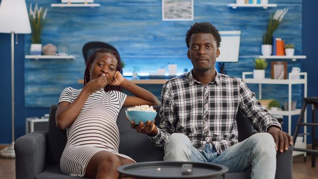 POV of black couple watching TV and expecting child in living room. African american pregnant woman laying on couch eating popcorn while man sitting, helping with pregnancy at home