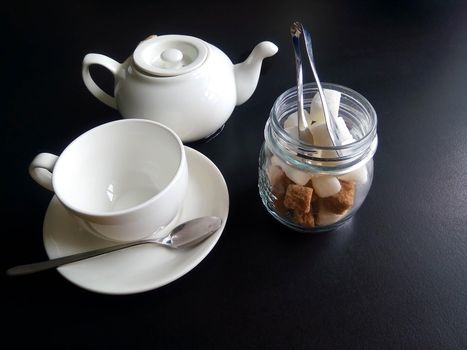 A white cup and saucer, a jar of sugar and a teapot, isolated on a black background.