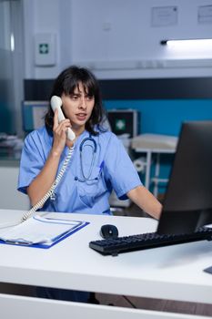 Medical assistant talking on telephone for checkup appointment, working late. Woman nurse using landline phone for online remote communication with patient at healthcare cabinet.