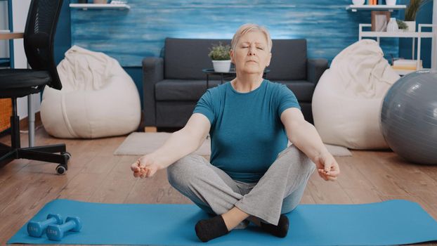 Retired woman with closed eyes sitting in lotus position to practice meditation on yoga mat. Old person meditating to find balance and harmony, relaxing with activity. Fitness training