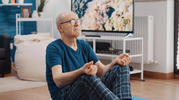 Handheld shot of pensioner in lotus position meditating and relaxing after fitness exercise. Senior man sitting on yoga mat and doing zen meditation for wellness and recreation.