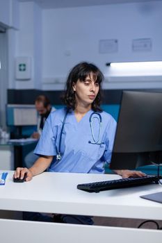 Medical assistant using computer at desk for work overtime. Nurse in uniform looking at monitor for paperwork and healthcare checkup, working late at office. Woman with expertise
