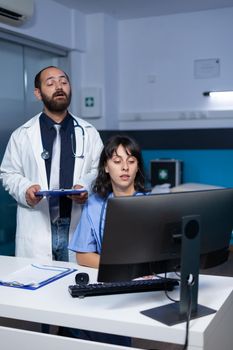 Doctor and nurse working with technology on computer for medical care and treatment. Team of specialists using monitor and examination files for healthcare, working late at night.