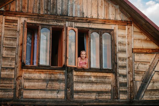 Silhouette of a young woman in the window of a wooden house in the mountains. Calm rest, alone with nature. In harmony with yourself. Meditation. Solitude in the mountains. Portrait of beautiful woman with long hair at the window frame of wooden, forest house