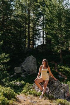 Pretty young girl in the yellow T-shirt and yellow shorts with sunglasses sitting on the stone above the beautiful canyon with an emerald lake on the sunset in Spain. Traveler in the mountains.