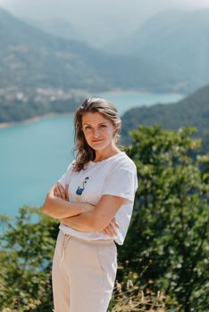 Girl tourist stands on the shore of a lake in the mountains. beautiful landscape, Piva Lake in Montenegro.. Woman is standing on the coast of Piva lake at sunset in summer. Landscape with girl, famous lake with beautiful reflection in water, trees, sky with clouds. Travel.