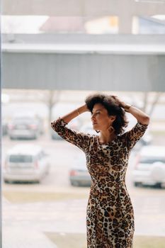 a brunette woman in a tiger dress stands near a large window.