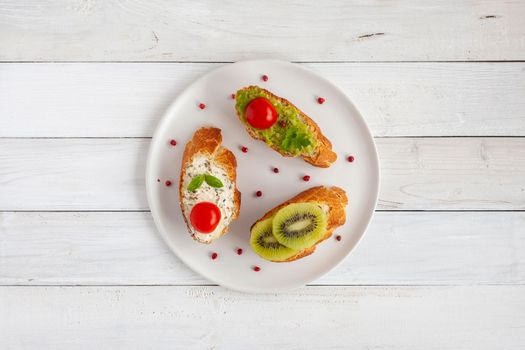 three homemade bright mini sandwichs with cream cheese and vegetables, on a white plate on wooden background, top view