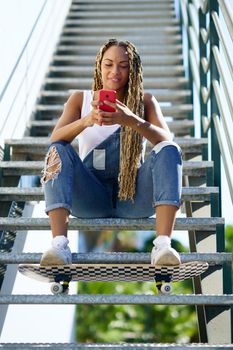 Young black woman with coloured braids, sitting on some steps while consulting her smartphone with her feet resting on a skateboard.