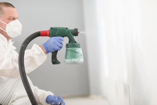 Portrait of worker using spray gun for coloring wall in white. Effective and fast way to paint walls. Renovation, interior design, household repair concept